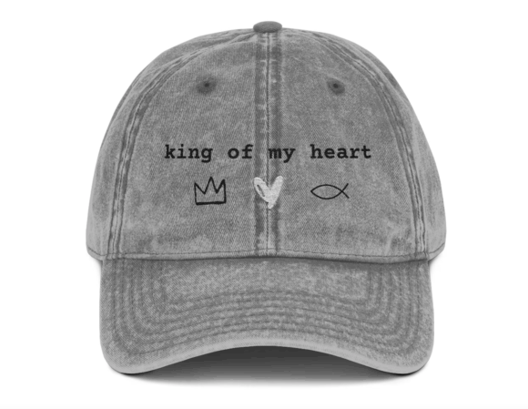 A gray, distressed baseball cap featuring the phrase "king of my heart" embroidered in black text across the front. Below the text are three simple symbols: a black crown, a white heart, and a black Christian fish symbol