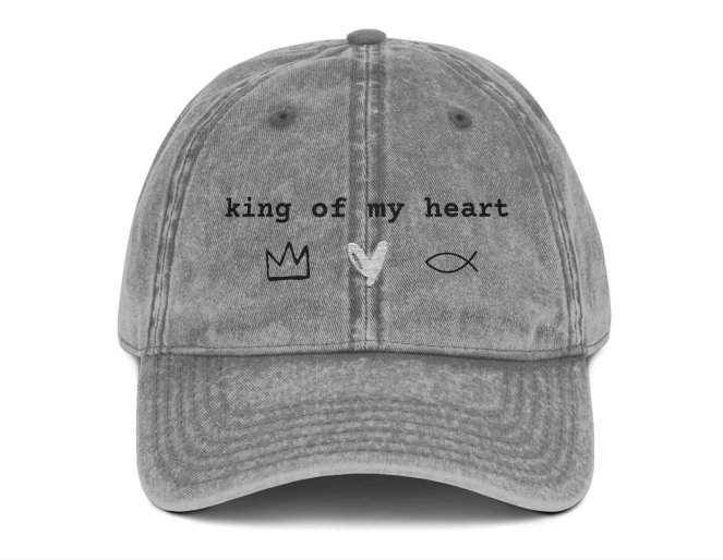 A gray, distressed baseball cap featuring the phrase "king of my heart" embroidered in black text across the front. Below the text are three simple symbols: a black crown, a white heart, and a black Christian fish symbol