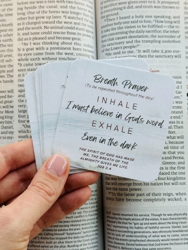 Hand holding breath praer cards set against an open Bible. The cards have simple, faith-based phrases for daily prayer and reflection.