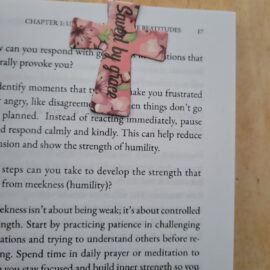 Cross-shaped magnetic bookmark with pink floral design clipped inside an open book, featuring an encouraging Christian phrase.