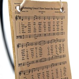 Rectangular ornament with an antique style hymnal page of "Amazing Grace" on it. The ornament is hanging by twine from a black ornament stand.