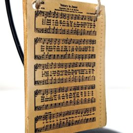 Rectangle shaped ornament with a yellowed hymnal page of "Victory in Jesus" on it. The ornament is hanging by twine from a black ornament stand.