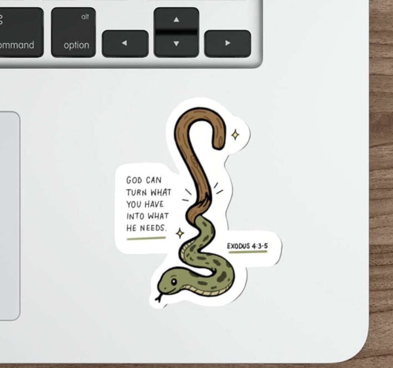 A die-cut sticker featuring a cartoon green and brown snake with the text 'GOD CAN TURN WHAT YOU DO INTO WHAT HE NEEDS' and 'EXODUS 4:2-4' on a white laptop palm rest, shown on a wooden desk next to a keyboard, mouse, and stylus.